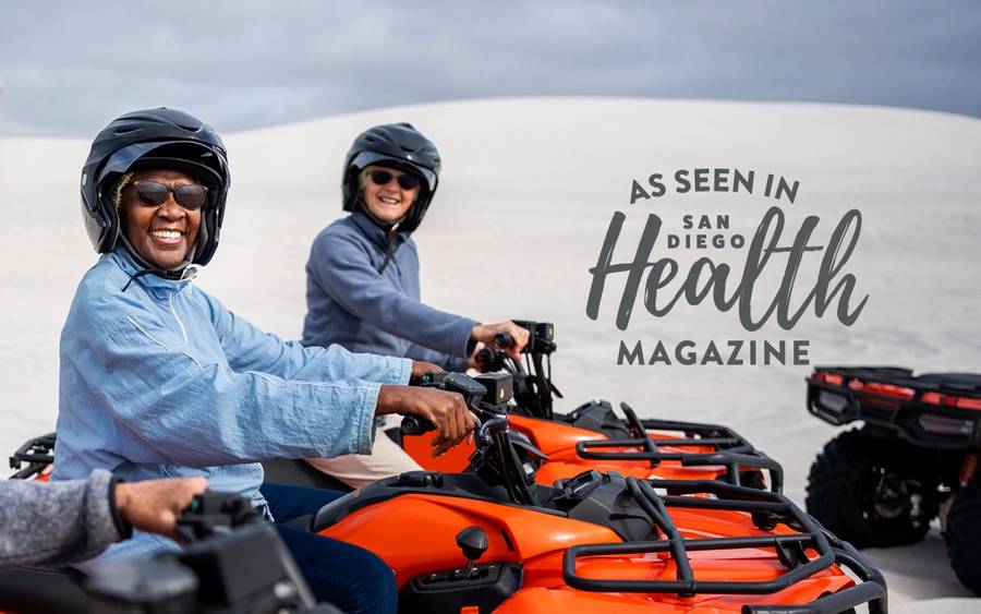 Two adults smile as they get ready to ride an ATV each wearing a helmet with a desert landscape in the background. SD Health Magazine