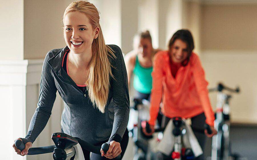 A trio of young women ride exercise bikes, representing Breast Cancer Awareness Month and info from a Scripps cancer expert.