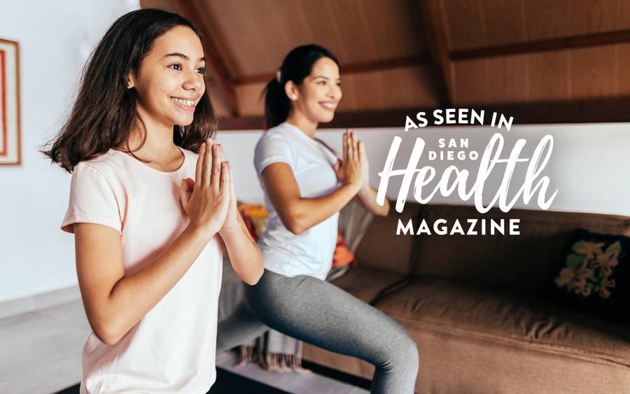 Two young women smile as they practice yoga on a mat indoors. SD Health Magazine