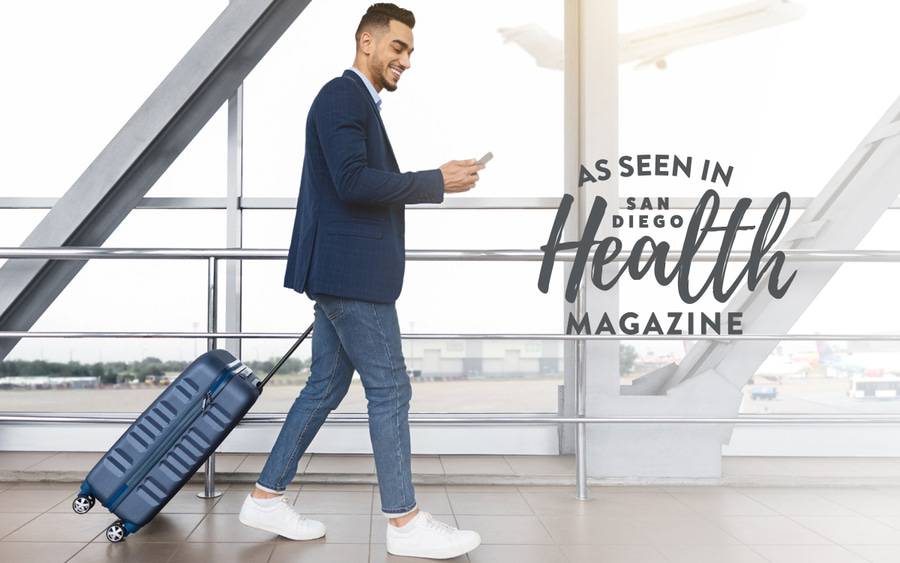 A young man smiles as he looks at his smartphone while walking through an airport terminal with a view of airplanes in the background. SD Health Magazine