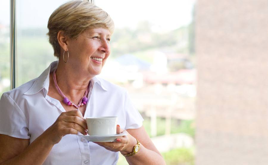 A smiling mature woman looks out the window with a cup of tea, representing breast cancer treatment at Scripps Cancer Center.