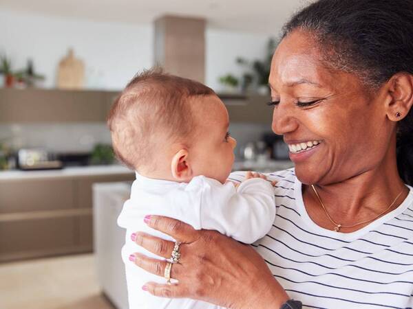 A mature woman in a kitchen smiles at a young infant in her arms. 