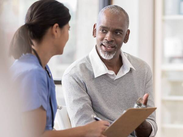 A mature male patient is discussing his health with a provider documenting notes on a clipboard illustrating the care provided by Ximed Medical Group providers.