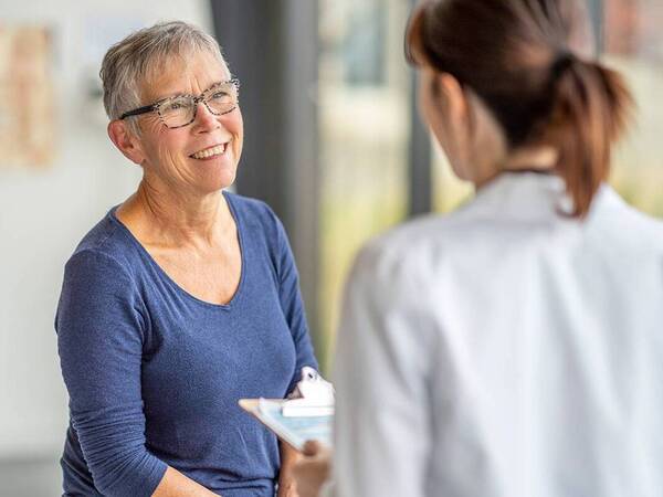 A mature woman wearing glasses is smiling and speaking with a health care provider.