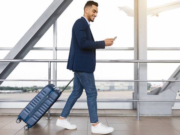 A young man smiles as he looks at his smartphone while walking through an airport terminal with a view of airplanes in the background.