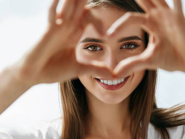 A young woman looks through her hands that are held up in the shape of a heart, representing the naturally beautiful appearance after cosmetic eye surgery.