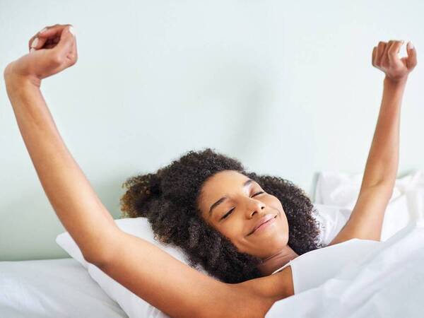 A woman smiles and stretches as she awakens in bed, feeling refreshed and alert after a good night’s sleep.