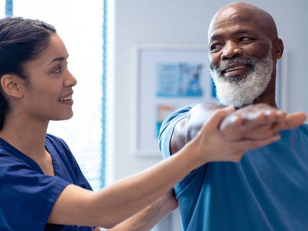 A physical therapist supports a patient's arm as he stretches his arm forward to help with joint stiffness from cancer treatment.