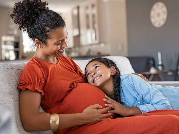 A pregnant woman in an orange dress sitting on the couch with her daughter and smiling.