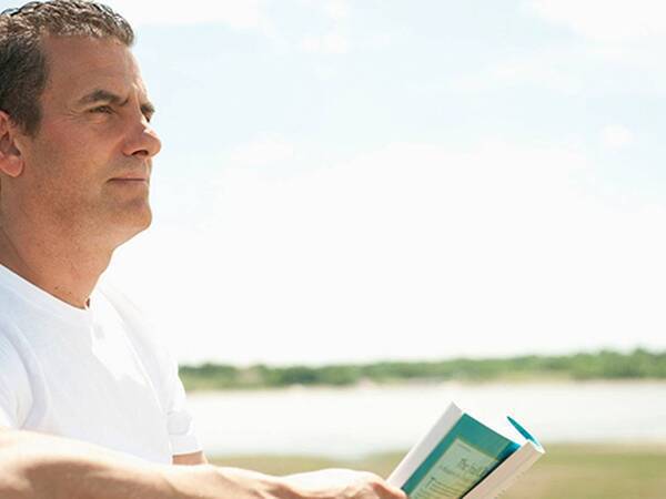 Middle-aged man reading a book near the shoreline.